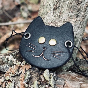 May include: A black leather cat charm with gold eyes and a stitched smile. The charm is attached to a black cord.