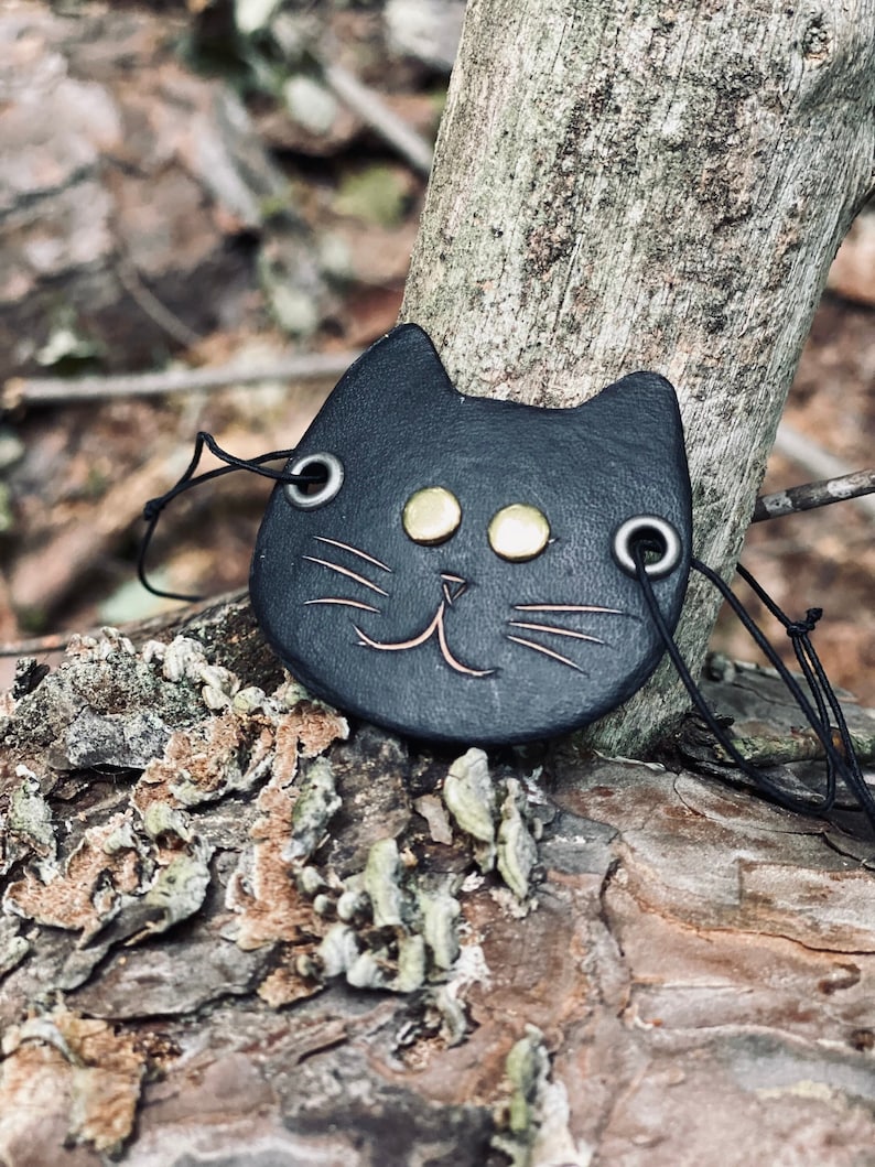 May include: A black leather cat charm with gold eyes and a stitched smile. The charm is attached to a black cord.