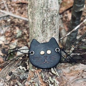 May include: Black leather cat charm with gold-colored eyes and whiskers. The charm is hanging from a black string and is resting on a tree trunk.