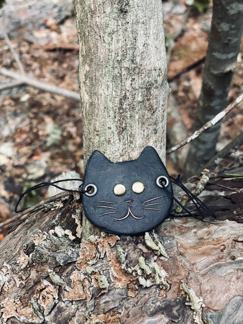 May include: Black leather cat charm with gold-colored eyes and whiskers. The charm is hanging from a black string and is resting on a tree trunk.