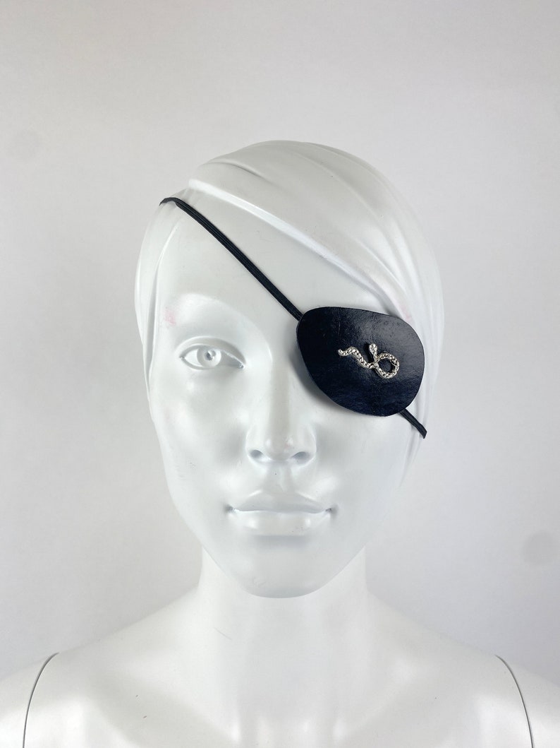 May include: A black leather eye patch with a rhinestone snake design. The eye patch is worn on a white mannequin head.