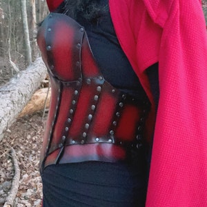 May include: A red leather corset with black metal accents and rivets. The corset is worn over a black top and a red hooded cloak.