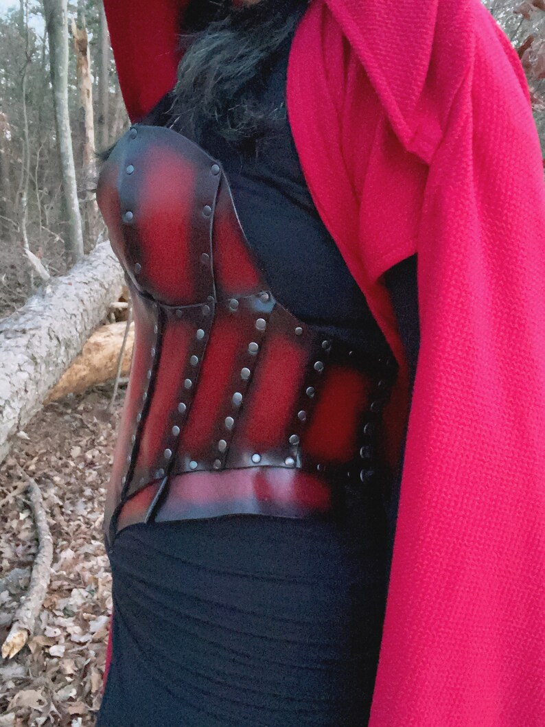 May include: A red leather corset with black metal accents and rivets. The corset is worn over a black top and a red hooded cloak.
