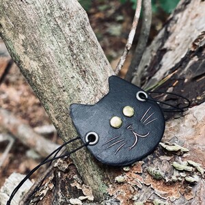 May include: Black leather cat charm with gold-colored eyes and a stitched smile. The charm is attached to a black cord.