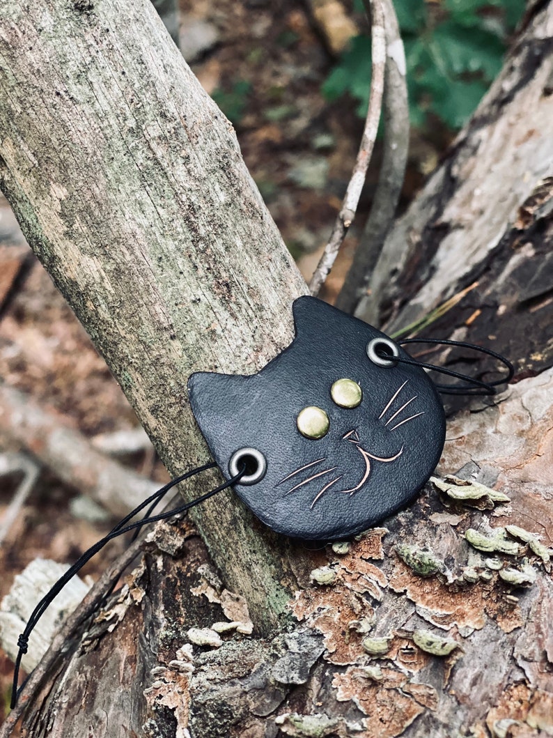 May include: Black leather cat charm with gold-colored eyes and a stitched smile. The charm is attached to a black cord.
