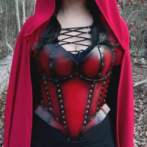May include: A red leather corset with black metal accents and a red hooded cape. The corset has a lace-up front and is decorated with rivets.