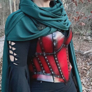 May include: A red leather corset with metal accents and rivets, worn over a black long-sleeved shirt and a green hooded cloak.
