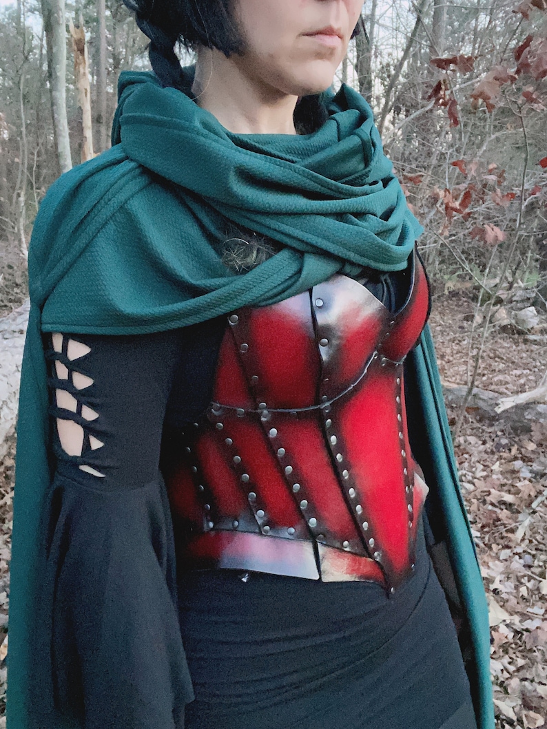 May include: A red leather corset with metal accents and rivets, worn over a black long-sleeved shirt and a green hooded cloak.