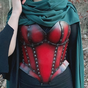 May include: A red leather corset with metal accents and rivets. The corset is worn over a black top and a green shawl.