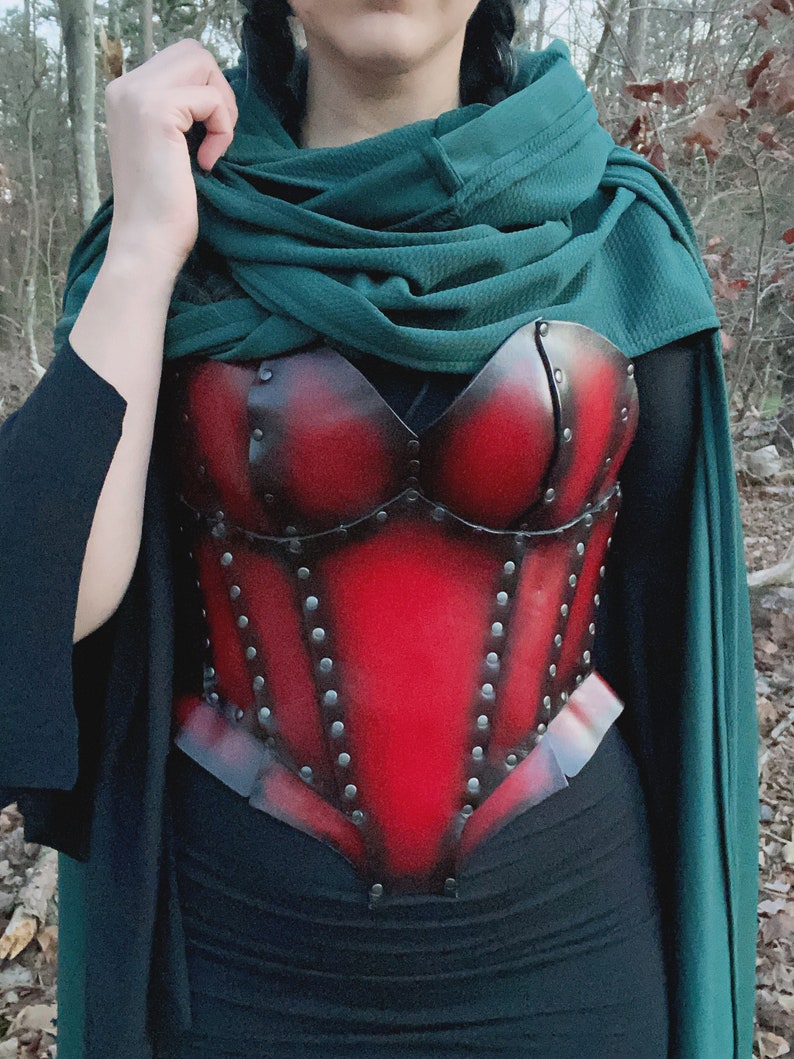 May include: A red leather corset with metal accents and rivets. The corset is worn over a black top and a green shawl.