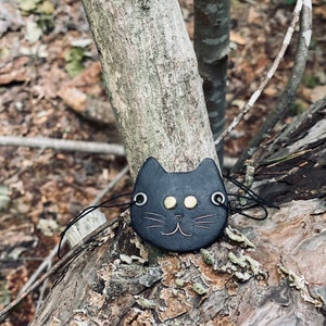 May include: A black leather cat charm with gold eyes and a stitched smile, hanging from a black cord on a tree branch.