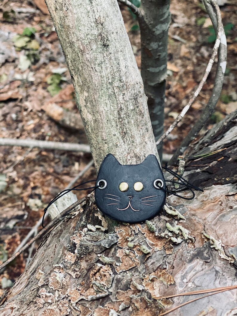 May include: A black leather cat charm with gold eyes and a stitched smile, hanging from a black cord on a tree branch.