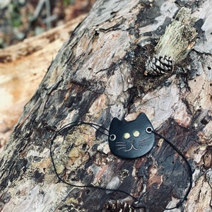 May include: Black leather cat face charm with gold eyes and a black cord. The charm is on a tree trunk with pine cones.