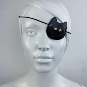 May include: A black leather eye patch with a cat face design. The patch has two gold-colored buttons for eyes and a stitched nose.