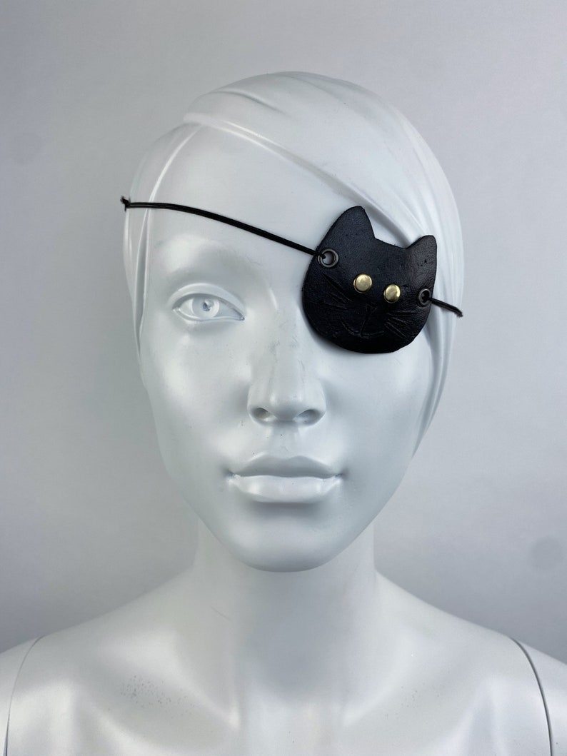 May include: A black leather eye patch with a cat face design. The patch has two gold-colored buttons for eyes and a stitched nose.