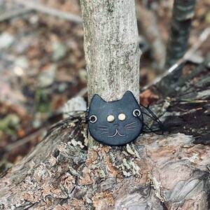 May include: A black leather cat charm with gold eyes and a stitched smile. The charm is hanging from a tree branch.