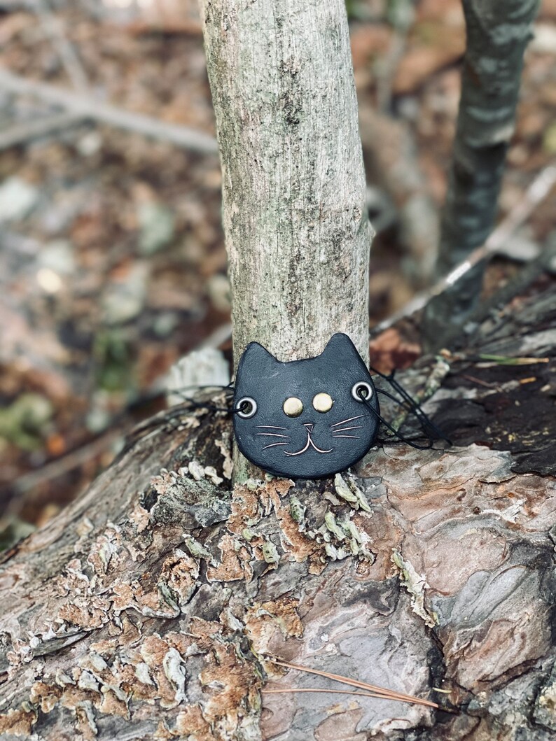 May include: A black leather cat charm with gold eyes and a stitched smile. The charm is hanging from a tree branch.