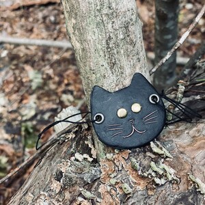 May include: A black leather cat charm with gold eyes and a stitched smile. The charm is attached to a black string and is hanging on a tree branch.