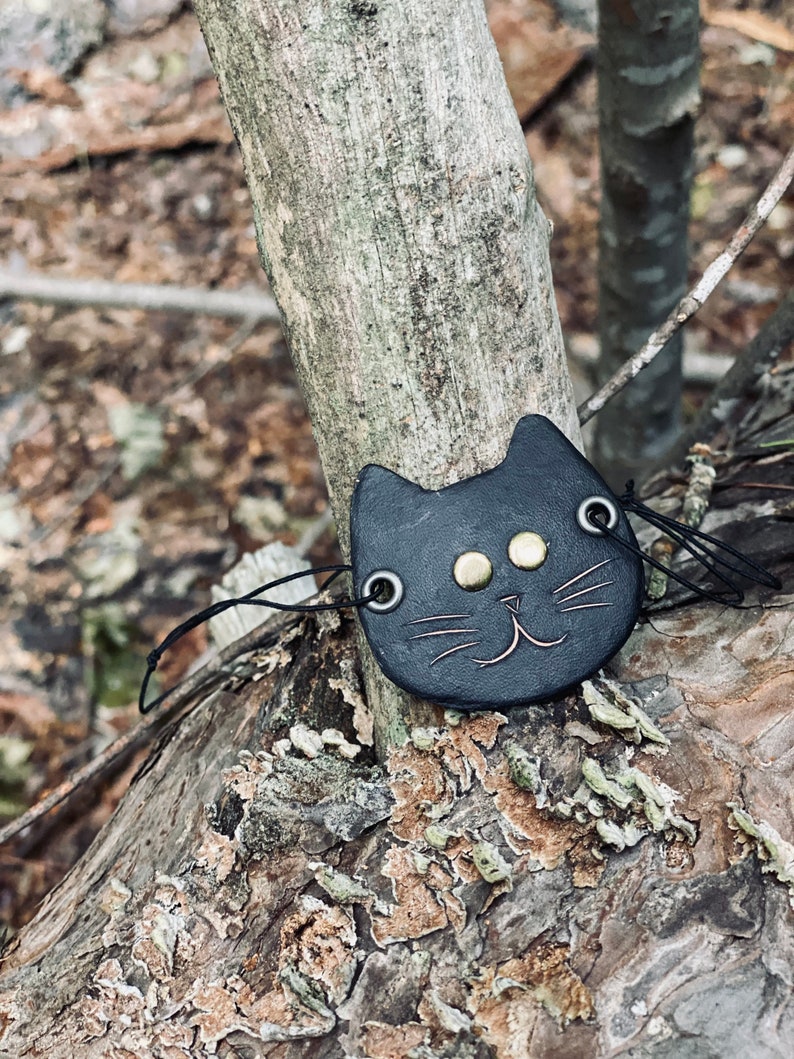 May include: A black leather cat charm with gold eyes and a stitched smile. The charm is attached to a black string and is hanging on a tree branch.