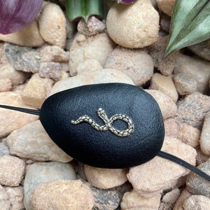 May include: A black leather eye patch with a silver snake design. The eye patch is on a bed of small, light brown rocks.
