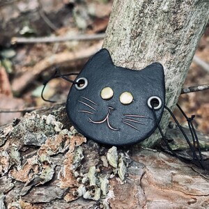 May include: Black leather cat charm with gold accents and a smiling face. The charm is hanging from a black cord.