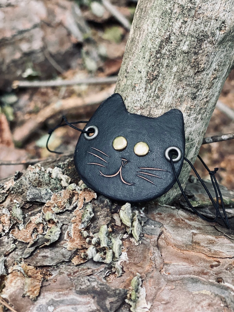 May include: Black leather cat charm with gold accents and a smiling face. The charm is hanging from a black cord.