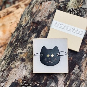 May include: A black leather cat face pin with gold eyes and a white background. The pin is in a small white box with the text "AGENT CRUSH CREATIVE" on the box.