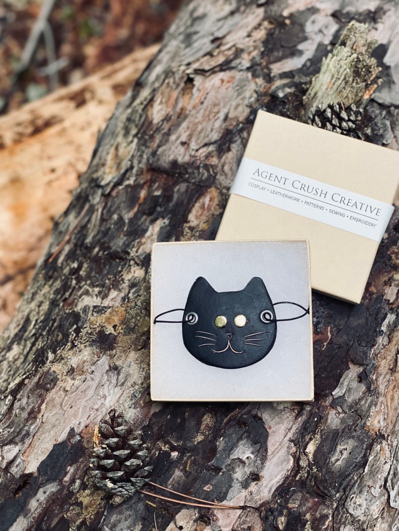 May include: A black leather cat face pin with gold eyes and a white background. The pin is in a small white box with the text "AGENT CRUSH CREATIVE" on the box.