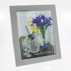 May include: A silver picture frame with a matted opening, holding a photo of a bouquet of purple irises in a crystal vase, a bottle of champagne in a crystal ice bucket, and two crystal glasses on a wooden table. The photo reads "Holds 8" x 10" Photo".