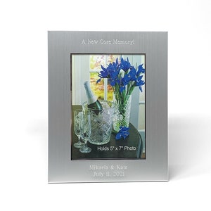 May include: Silver photo frame with a brushed finish. The frame holds a 5x7 inch photo of a bouquet of blue irises in a vase, a bottle of champagne in an ice bucket, and two champagne flutes. The frame is engraved with the text "A New Core Memory!" and "Holds 5" x 7" Photo". Below the photo is the text "Mikaela & Kate July 11, 2021".