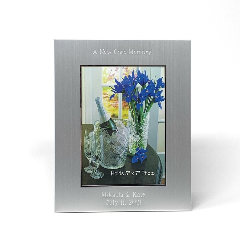 May include: Silver photo frame with a brushed finish. The frame holds a 5x7 inch photo of a bouquet of blue irises in a vase, a bottle of champagne in an ice bucket, and two champagne flutes. The frame is engraved with the text "A New Core Memory!" and "Holds 5" x 7" Photo". Below the photo is the text "Mikaela & Kate July 11, 2021".