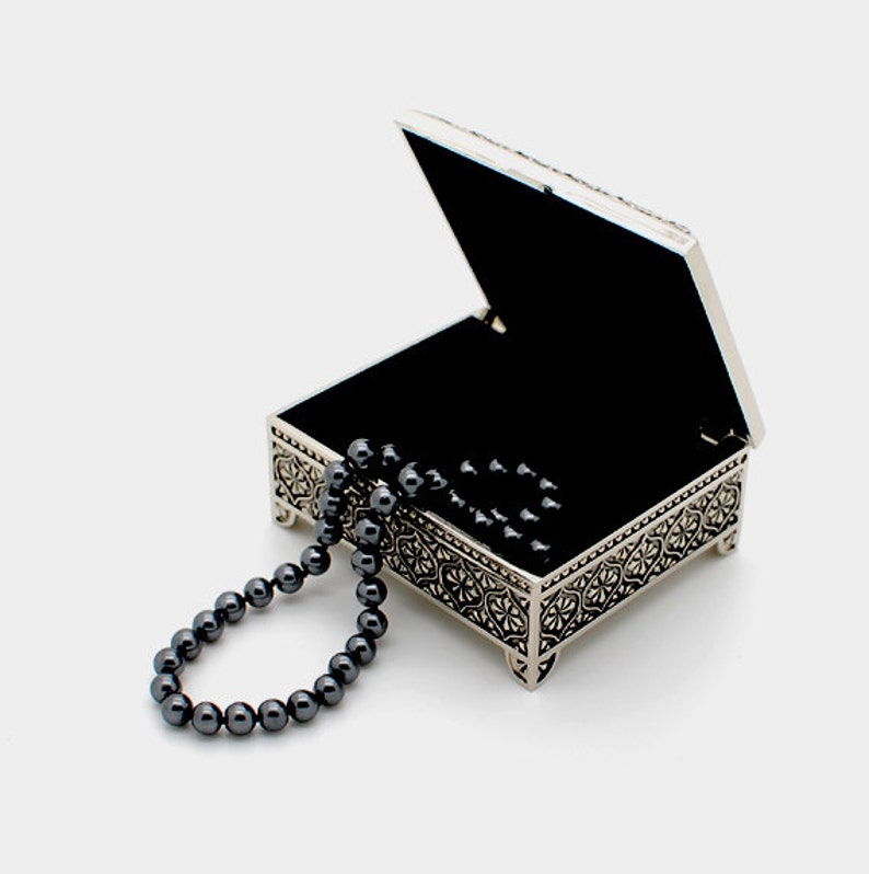 May include: An open silver metal trinket box with an ornate design and a black velvet interior. A black beaded necklace is lying on the box and spilling out of the box.