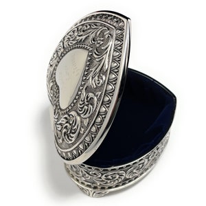May include: Silver heart-shaped trinket box with an ornate design and a blue velvet interior. The lid is open and the box is sitting on a white surface.