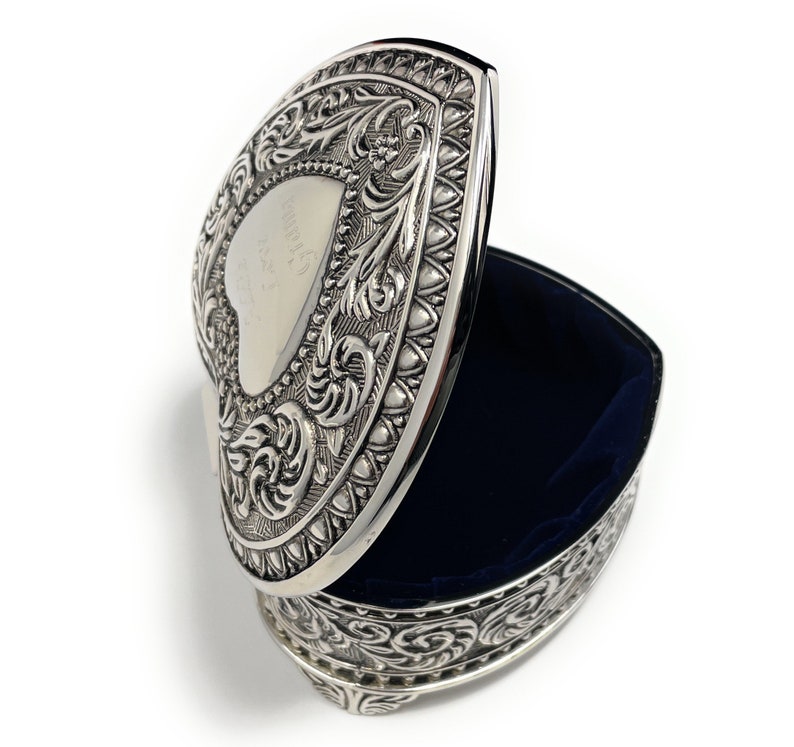 May include: Silver heart-shaped trinket box with an ornate design and a blue velvet interior. The lid is open and the box is sitting on a white surface.