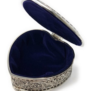 May include: A silver heart-shaped trinket box with an ornate design and a blue velvet interior. The lid is open, revealing the empty interior.