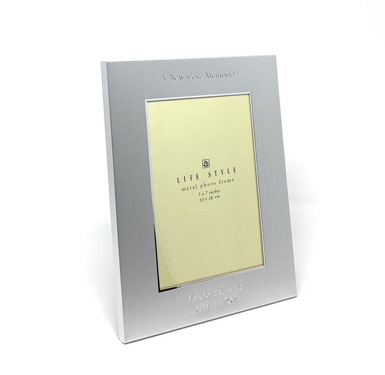 May include: A silver metal photo frame with a 5x7 inch opening. The frame has the text "A New Core Memory" and "LIFE STYLE metal photo frame 5x7 inches 13x18 cm" engraved on the front. The frame is personalised with the text "Mikasa & Rose July 4, 2021".