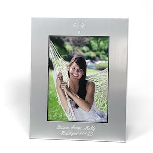 May include: Silver picture frame with a cross design and a photo of a person in a hammock. The frame has the text "Saxon James Kelly Baptized 11120" engraved on the bottom.