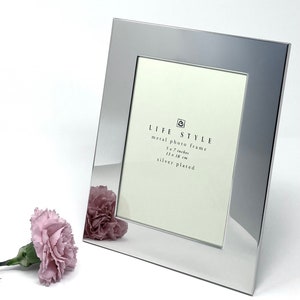 May include: A silver-plated metal photo frame with a 5x7 inch opening. The frame is labeled "LIFE STYLE" and has the dimensions 13x18 cm.