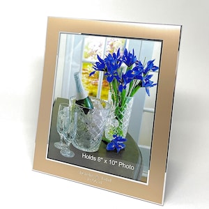 May include: A gold picture frame with a silver border holds an 8x10 inch photo of blue irises in a vase and a bottle of champagne in a crystal ice bucket. The frame has an inscription that reads "Holds 8" x 10" Photo" and "Don & Tabi 12.06.21".