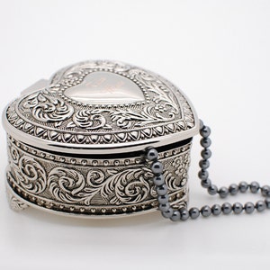 May include: A silver heart-shaped trinket box with an ornate design and a beaded chain.