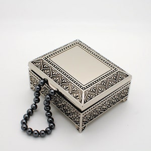 May include: A silver metal trinket box with an ornate design and a black beaded chain handle.