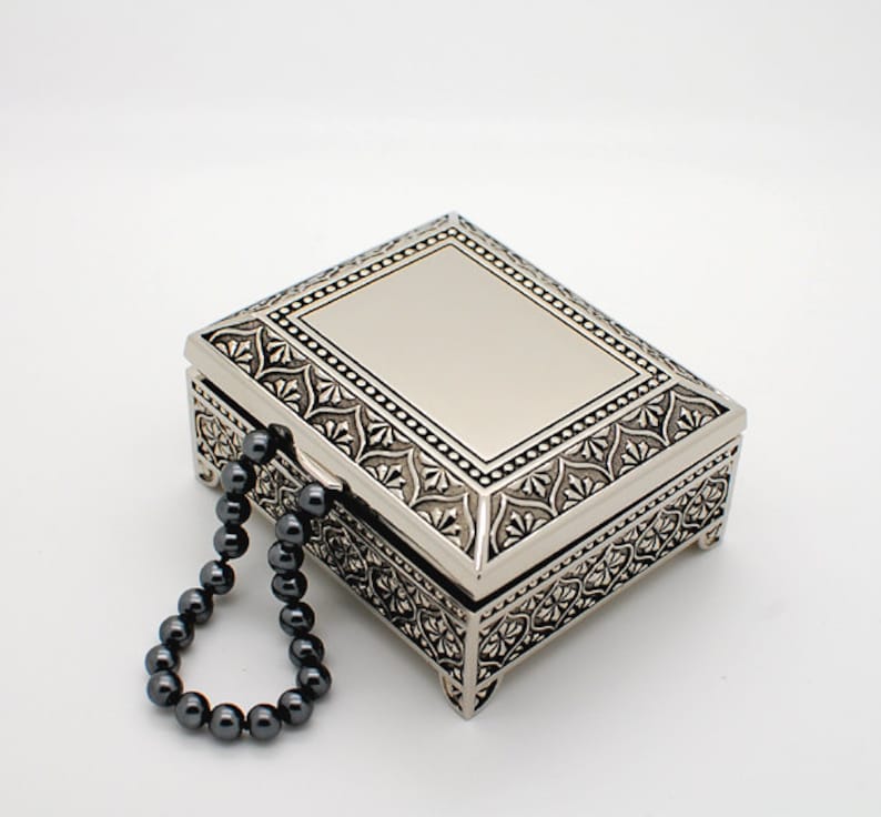 May include: A silver metal trinket box with an ornate design and a black beaded chain handle.
