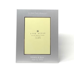 May include: Silver metal photo frame with a 5x7 inch opening. The frame has a brushed finish and is engraved with the text "A New Core Memory!" and "LIFE STYLE metal photo frame 5 x 7 inches 13 x 18 cm". The frame also has a personalized inscription that reads "Mikaela & Kate July 11, 2021".