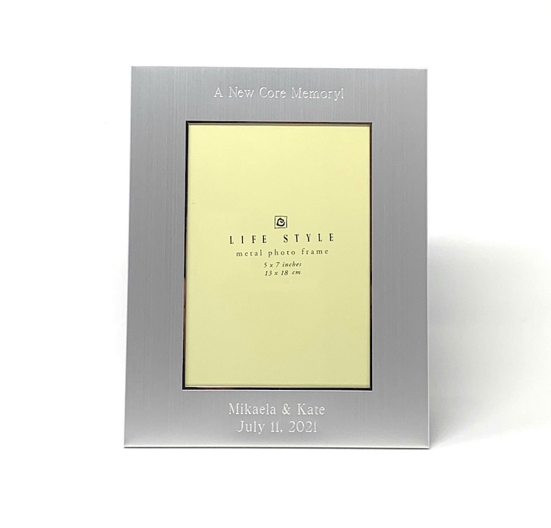 May include: Silver metal photo frame with a 5x7 inch opening. The frame has a brushed finish and is engraved with the text "A New Core Memory!" and "LIFE STYLE metal photo frame 5 x 7 inches 13 x 18 cm". The frame also has a personalised inscription that reads "Mikaela & Kate July 11, 2021".