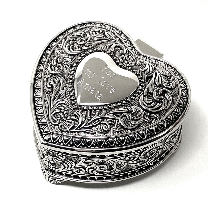 May include: Silver heart-shaped trinket box with an ornate design and a hinged lid. The lid features an engraved heart with the inscription "For my love Amata".