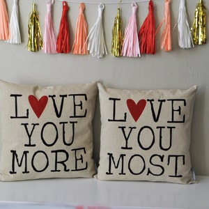May include: Two beige throw pillows with the text "LOVE YOU MORE" and "LOVE YOU MOST" printed in black with a red heart in the middle of each pillow.