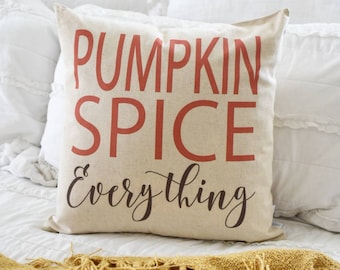May include: A beige square pillow with the words "PUMPKIN SPICE Everything" printed in orange and black. The pillow is on a white bed with a yellow fringed blanket.