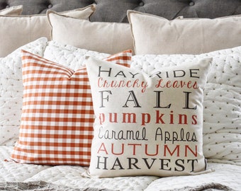 May include: Two decorative throw pillows on a bed. One pillow has a red and white gingham pattern. The other pillow is beige with the words "Hay Ride, Crunchy Leaves, Fall, Pumpkins, Caramel Apples, Autumn, Harvest" in black and red.