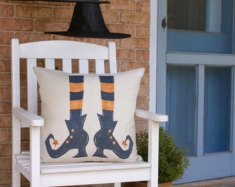 May include: A white wooden chair with a decorative pillow featuring witch legs in blue and orange stripes. Black witch hats hang above. A carved pumpkin sits on the porch, suggesting a Halloween theme.