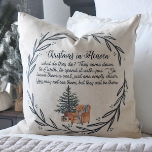May include: A beige throw pillow with the text "Christmas in Heaven" and a poem. The design includes a Christmas tree and a wreath of leaves. The pillow is on a bed with white bedding.